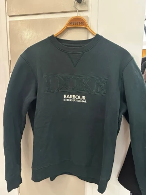 Mörkgrön sweatshirt Barbour International - Snygg mörkgrön sweatshirt från Barbour International i storlek M. Tröjan har broderad DUKE-text på bröstet och tryckt logga under. Rund hals och långa ärmar, tillverkad i mjuk bomull. Perfekt för chill och streetwear.