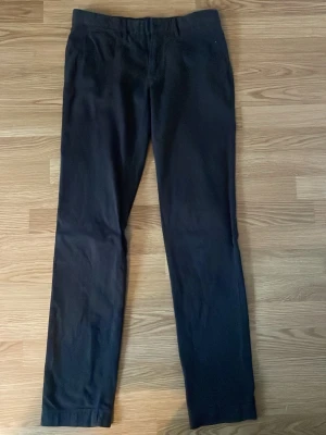 Mörkbruna kostymbyxor J.Lindeberg - Snygga svarta kostymbyxor från J.Lindeberg i storlek 30/34. Klassisk rak passform med två bakfickor med knapp och bälteshällor. Perfekta för en clean och stilren look. Materialet är mjukt och känns lyxigt mot huden.