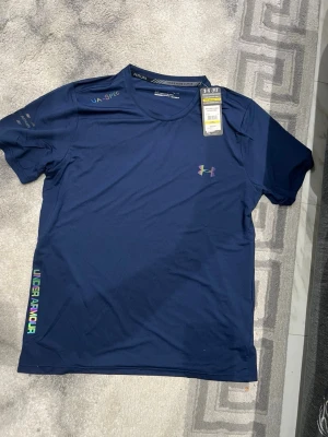 Blå Under Armour tränings-tshirt - Snygg marinblå tränings-tshirt från Under Armour Tillverkad i lätt och ventilerande syntetmaterial, perfekt för träning. Logga och text i regnbågsfärger på bröstet och sidan. Rund hals och kort ärm för maximal rörelsefrihet. Osäker på storleken men den är ganska liten i storlek, den sitter dessutom slim fit så den är som en compression T-shirt  priset kan diskuteras 😊