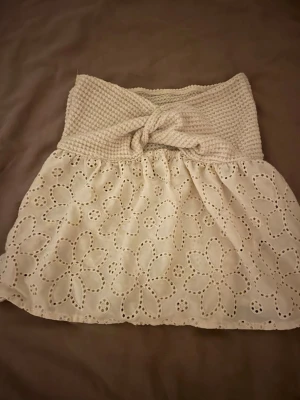 Beige tubtopp med spets och knyt - Säljer en beige tubtopp med virkad överdel och knyt framtill. Nederdelen är i bomull med broderade blommor och spetsdetaljer. Perfekt för sommarens varma dagar och har en söt, feminin look med bandeau-stil.
