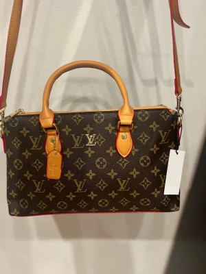 Louis Vuitton monogram axelväska - Säljer en snygg axelväska från Louis Vuitton med klassiskt monogrammönster i brunt och beige. Väskan har både handtag och justerbar axelrem i ljusbrunt skinn med röda detaljer. Guldiga metalldetaljer och dragkedja upptill. Perfekt storlek för dina essentials.