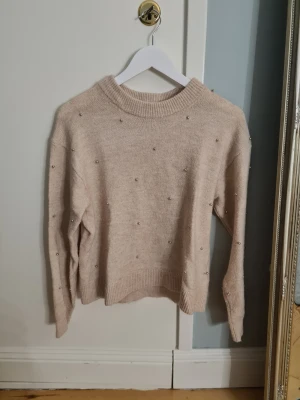 Beige stickad tröja med pärlor från H&M - Säljer en beige stickad tröja från H&M i storlek XS. Tröjan har rund hals, ribbade muddar och är dekorerad med små pärlor över hela framsidan. Perfekt för dig som gillar enkla detaljer och mjuka material.