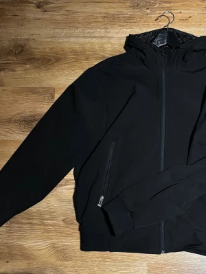 Svart vindjacka från Stone Island - Säljer en svart vindjacka från Stone Island med huva och dragkedja framtill. Jackan har en klassisk patch på ärmen och praktiska fickor med dragkedja. Materialet är polyester och jackan har en clean, stilren look som passar perfekt till streetwear.