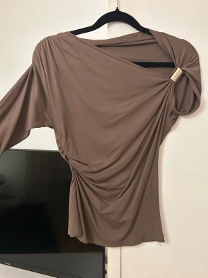 Zara topp - Säljer en brun oneshoulder topp i mjukt stretchigt material. Toppen har en snygg ringdetalj i guld på axeln och draperad effekt över kroppen. Passformen är slim och toppen är perfekt för dig som gillar trendiga och unika plagg.