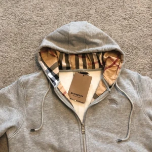 Grå hoodie från Burberry med dragkedja - Säljer en grå hoodie från Burberry i storlek L. Klassisk dragkedja framtill, huva med ikoniskt rutigt foder och snörning. Broderad logga på bröstet. Perfekt för dig som vill ha en stilren och lyxig vardagslook.