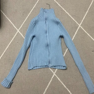 Blå ribbad kofta med dragkedja - Säljer en ljusblå ribbad kofta med hög krage och hel dragkedja framtill. Tröjan är långärmad och har en tight passform med stretchigt stickat material. Perfekt för lager-på-lager under kyliga dagar.