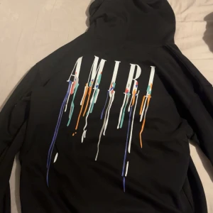 Svart Amiri hoodie med färgstänk - Svart hoodie från Amiri med stor logga och färgstänk på ryggen samt mindre tryck på bröstet. Klassisk huva med snörning och känguruficka framtill. Snygg streetwear-vibe och riktigt skön passform. Passar vikterna från 77kg - 95kg 