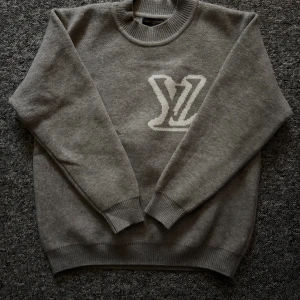Grå stickad Louis Vuitton tröja M - Snygg grå stickad tröja från Louis Vuitton med stort vitt LV-logo på både fram- och baksidan. Tröjan har rund hals, ribbade muddar och är långärmad. Perfekt för dig som gillar exklusiva märken och vill ha en clean look. Passar bra till någon som är mellan 185-190cm lång