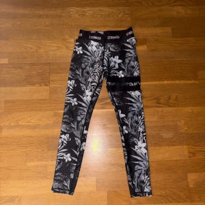 Stronger Pompeii leggings med blommigt mönster - Snygga svarta leggings från Stronger i modellen Pompeii med grått och vitt blommigt mönster. Hög midja och bred resår med logga. Tillverkade i stretchigt material av polyester och spandex, perfekta för träning eller chill.