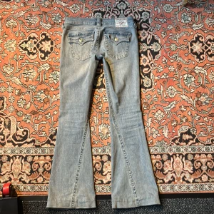 True Religion Joey Low Rise Flare Jeans - Snygga ljusblå jeans från True Religion, modell Joey Low Rise Flare. Byxorna har låg midja, utsvängda ben och klassiska detaljer som markerade sömmar och fickor med  knappar. Perfekta för dig som gillar en retroinspirerad look. OBS! Jeansen är uppsydda i benen 5 cm då de var för långa när jag köpte dem! Jag är 170 själv