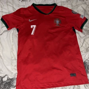 Portugal Ronaldo 7 Nike fotbollströja - Portugal landslagströja med nummer 7 och Ronaldo på ryggen. Röd med gröna och svarta detaljer vid krage och ärmslut, broderade märken på ärmarna och Nike-logga på bröstet. Tillverkad i lätt och ventilerande polyester, perfekt för match eller träning.