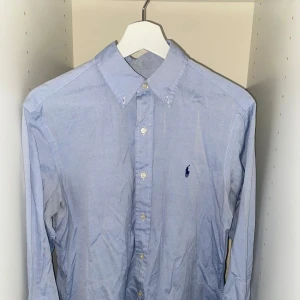 Blå skjorta från Ralph Lauren - Klassisk ljusblå skjorta från Ralph Lauren. Tillverkad i 100% bomull och har en normal passform (Classic Fit). Skjortan är i storlek 15 32/33 vilket passar storlek m. Vid fler frågor skicka ett meddelande.