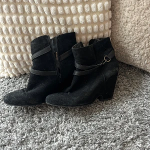 Svarta mockaboots med klack - Snygga svarta boots i mocka med rund tå och hög klack. Skorna har dekorativa remmar med metallspänne runt ankeln och dragkedja på sidan för enkel påtagning. Perfekt för dig som gillar stilrena och trendiga skor med lite edge.