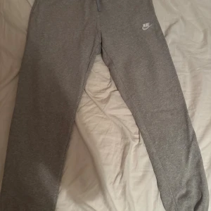 Grå mjukisbyxor från Nike - Grå mjukisbyxor från Nike med klassisk logga på vänster lår. Byxorna har elastisk midja med snörning och ribbade muddar vid bensluten. Tillverkade i mjukt bomullsmaterial för maximal komfort. Perfekta för chill eller träning.