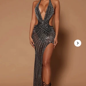 Glittrig svart långklänning med slits - Säljer en svart långklänning med glittriga detaljer och djup v-ringning. Klänningen har en hög slits på ena sidan, öppna partier vid midjan och är ärmlös med halterneck. Perfekt för dig som vill sticka ut på bal eller fest!