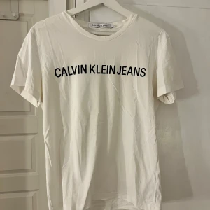 Vit t-shirt från Calvin Klein Jeans - Vit t-shirt från Calvin Klein Jeans med svart logotyptryck över bröstet. Klassisk rund halsringning och korta ärmar. Tillverkad i mjuk bomull för en skön och avslappnad känsla.