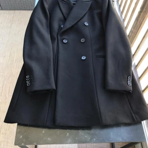 Svart rock från J.Lindeberg, stl 40 - Stilren svart rock från J.Lindeberg med dubbelknäppning och klassisk krage. Jackan har släta fickor, fyra knappar vid ärmslut och är helfodrad. Perfekt för dig som vill ha en tidlös och snygg look under kallare månader.