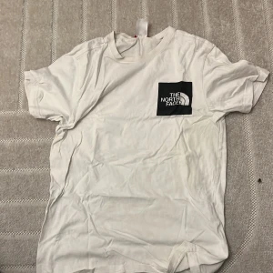 Vit t-shirt från The North Face - Snygg vit t-shirt från The North Face med svart logga på bröstet och mindre logga på ryggen. Klassisk passform och rund halsringning. Tillverkad i mjuk bomull som är skön mot huden. Perfekt för en avslappnad och stilren look.