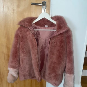 Rosa pälsjacka från Lindex, stl 152 - Supermjuk och fluffig pälsjacka från Lindex i en snygg rosa nyans med ljusare detaljer vid ärmsluten. Jackan har dragkedja framtill och är helfodrad för extra komfort. Perfekt för dig som vill sticka ut med en cozy vibe.