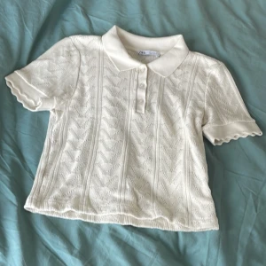 Vit stickad blus med krage från Zara - Supersöt vit stickad blus från Zara med klassisk krage och korta ärmar. Blusen har ett diskret zigzag-mönster, tre knappar framtill och vågiga kanter på ärmarna. Materialet är en mjuk mix av viskos och polyester som känns skönt mot huden. Storlek står S men känns som XS! 