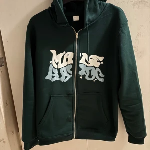 Mörkgrön hoodie med tryck och dragkedja - Mörkgrön hoodie med coolt vitt och ljusblått tryck på bröstet, huvan och ärmen. Tröjan har dragkedja framtill, kängurufickor och justerbar huva. Perfekt för dig som gillar streetstyle och vill sticka ut lite extra.