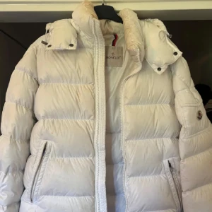Vit Moncler dunjacka med huva - Säljer en snygg vit dunjacka från Moncler med avtagbar huva och klassisk logga på ärmen. Jackan har dragkedja framtill, två fickor med dragkedja och quiltad design. Perfekt för kalla dagar och riktigt stilren att bära till allt.