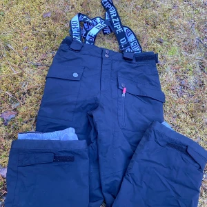 Svarta täckbyxor från Dr. Zipe - Svarta täckbyxor från Dr. Zipe med justerbara hängslen med logga, stora fickor med knappar och dragkedja, samt kardborre vid benslut. Byxorna har en loose passform och är fodrade för extra värme. Perfekta för kalla dagar utomhus.