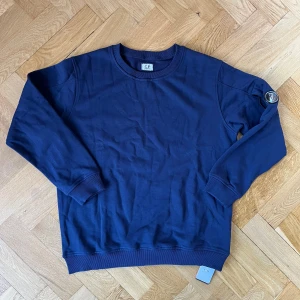 Mörkblå sweatshirt från C.P. Company - Snygg mörkblå sweatshirt från C.P. Company med klassisk rund hals och ribbade muddar. Tröjan har den ikoniska linsen på ärmen och är gjord i mjuk bomull. Perfekt för dig som gillar stilrena och sportiga plagg med streetkänsla.