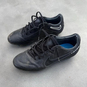 Nike Tiempo svarta fotbollsskor - Svarta Nike Tiempo fotbollsskor med klassisk Swoosh-logga i vitt och blå detaljer på sulan. Skorna har snörning, rund tå och ovandel i skinn för bra känsla på planen. Perfekta för dig som vill ha stilrena och funktionella skor till match eller träning.