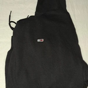 Svart hoodie från Tommy Jeans XL - Svart hoodie från Tommy Jeans i storlek XL. Klassisk design med liten broderad logga på bröstet, huva med snörning och stor magficka. Mjuk insida och ribbade muddar. Perfekt för dig som gillar streetwear och enkel stil.