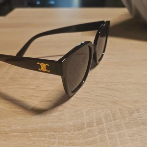 Svarta solglasögon med guldlogga - Snygga svarta solglasögon med en cool, kantig form och mörka linser. På skalmen finns en elegant guldfärgad logga som ger extra lyxig känsla. Glasögonen är tillverkade i blank syntet och passar perfekt för dig som gillar statement-accessoarer.