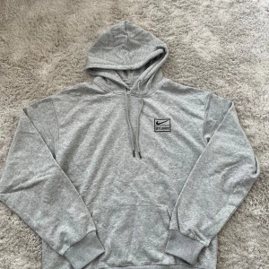 Grå Nike x Stussy hoodie - Säljer en grå hoodie från Nike i samarbete med Stussy. Tröjan har huva med snörning, magficka och broderad logga på bröstet. Tillverkad i mjuk bomullsmix och har en klassisk loose passform med långa ärmar. Perfekt för chill dagar eller streetwear-stil.