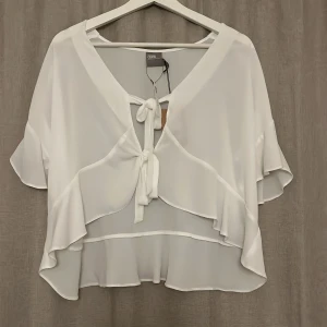 Vit omlottblus med volang från ASOS - Superfin vit blus från ASOS med omlottdesign och knyt framtill. Blusen har volangdetaljer längs ärmar och nederkant, samt en luftig och flowy passform. Perfekt för dig som gillar romantiska och trendiga plagg. 