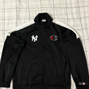 Svart vindjacka Champion x Yankees L - Svart vindjacka från Champion i samarbete med New York Yankees. Jackan har vita detaljer längs axlarna, broderade loggor på bröstet och elastiska muddar. Materialet är 100% polyester och jackan har hel dragkedja framtill. Perfekt för dig som gillar sportig streetstyle.