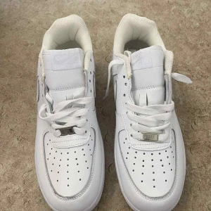 Nike Air Force 1 helvita sneakers - Fräscha Nike Air Force 1 sneakers i helvitt med klassisk låg siluett. Skorna har perforerad tå, snörning med metallbricka och tydlig Nike-logga bak. Tillverkade i skinn med platt sula och rund tå. Perfekta för dig som gillar cleana och stilrena sneakers.