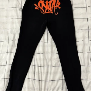 Svarta mjukisbyxor från SYNA med tryck - Svarta mjukisbyxor från SYNA med orange graffiti-tryck bak och broderad text 'Forgive Me' vid benet. Byxorna har snörning i midjan och dragkedja vid benslut. Materialet är 80% bomull och 20% polyester, vilket gör dem mjuka och sköna.