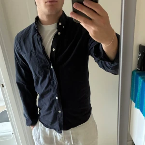 Mörkblå skjorta från ASKET - Snygg mörkblå skjorta från ASKET med klassisk button-down krage och vita knappar. Skjortan har en bröstficka och är tillverkad i mjuk bomull. Passformen är normal och ärmarna är långärmade. Perfekt för dig som gillar stilrena och enkla plagg. Lätt urtvättad men skapar en ny färg så funkar bra.