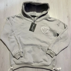 Grå  Carrsicko hoodie - Grå hoodie från Carrsicko med en vit broderad hjärtdetalj på bröstet. Tröjan har huva, ribbade muddar och snörning i nederkant. Mjuk insida och klassisk känguruficka framtill. Perfekt för chill dagar och streetwear.