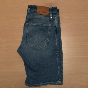 Blå Replay jeansshorts - Säljer ett par blå jeansshorts från Replay, modell RBJ.901 Short Tapered Fit. Klassisk femficksdesign, slitna detaljer framtill och knappgylf. Snyggt läderpatch bak i midjan. Perfekta för en avslappnad stil i sommar.