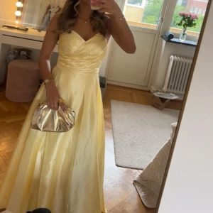 Ljusgul balklänning !!! - Säljer en elegant ljusgul långklänning i glansigt satinmaterial. Klänningen har omlottdrapering över bysten, v-ringad urringning och öppen rygg med snygg snörning som korsas och knyts baktill. Perfekt för bal eller student! Passar dig som vill sticka ut med en chic och feminin look.