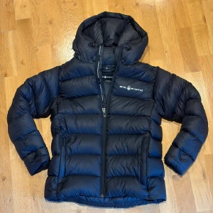 Svart dunjacka från Sail Racing - Säljer en svart, fluffig dunjacka från Sail Racing med huva och dragkedja. Jackan har en clean look med logga på bröstet och praktiska fickor med dragkedja. Perfekt för kalla dagar när du vill hålla stilen och värmen. Jackan har lite smått läckage på dun men absolut inget som förstör. Skriv om du har frågor.