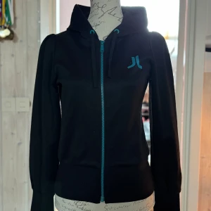 Svart hoodie från WESC med blå detaljer - Svart hoodie från WESC med blå dragkedja och logga på bröstet. Tröjan har huva med snörning och långa ärmar. Perfekt för dig som gillar streetwear och vill ha en stilren look med en färgklick.