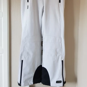 Vita skidbyxor från Killtec - Snygga vita täckbyxor från Killtec med svarta detaljer och dragkedjor vid bensluten. Byxorna har praktiska fickor med dragkedja och snygga sömmar över knä. Perfekta för kalla dagar, slalom och vinteraktiviteter.