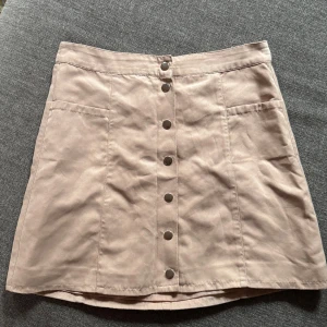 Beige kortkjol med knappar från H&M - Snygg beige kortkjol från H&M Divided i storlek 38. Kjolen har raka linjer, stora fickor framtill och knappar hela vägen ner. Perfekt för dig som gillar en enkel men trendig look. Materialet känns mjukt och har en lätt struktur.