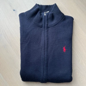 Svart stickad kofta från Ralph Lauren - Svart stickad kofta från Ralph Lauren med röd broderad logga på bröstet. Tröjan har ribbad hög krage, hel dragkedja framtill och långa ärmar. Perfekt att slänga på sig över en t-shirt för en clean och klassisk look.
