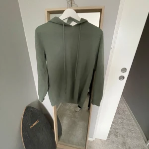 Olivgrön hoodie med huva - Säljer en olivgrön hoodie med huva och dragsnören. Tröjan är långärmad och har en enkel, clean look utan tryck eller mönster. Materialet är merinoull som känns mjukt och lite stickat, perfekt för kyligare dagar. Passformen är normal och den funkar till det mesta.
