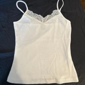 Vit ribbad linnetopp med spets - Säljer en vit ribbad linnetopp med smala axelband och v-ringning dekorerad med spets. Perfekt för sommardagar eller som lager under en skjorta. Enkel och stilren design som passar till det mesta.