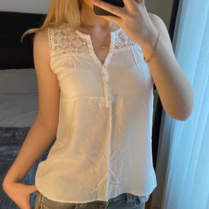 Vit ärmlös blus med spets från Soaked - Supersöt vit blus från Soaked med ärmlös design och spetsdetaljer upptill. Blusen har knappar framtill och ett luftigt, lätt material som passar perfekt till jeans eller kjol. Spetsen har rutigt mönster och ger en romantisk vibe. Perfekt för varma dagar!