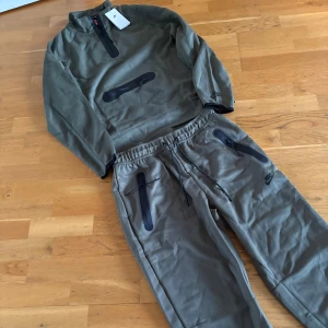 Nike gröna mjukisbyxor med dragkedja - Snygga gröna mjukisbyxor från Nike med svarta detaljer och dragkedjor på fickorna. Byxorna har snörning i midjan och en loose passform som är perfekt för chill eller träning. Materialet är mjukt och skönt, och Nike-loggan syns på benet.