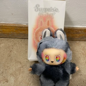 Pop Mart Surprise Baby Pretty - Pop Mart Surprise Baby Pretty figur med originalförpackning. Söt samlarfigur i mjuk grå kanindräkt, färgglada detaljer och regnbågsögon. Mycket fint skick, inga synliga defekter eller slitage. Perfekt för samlare eller som present.